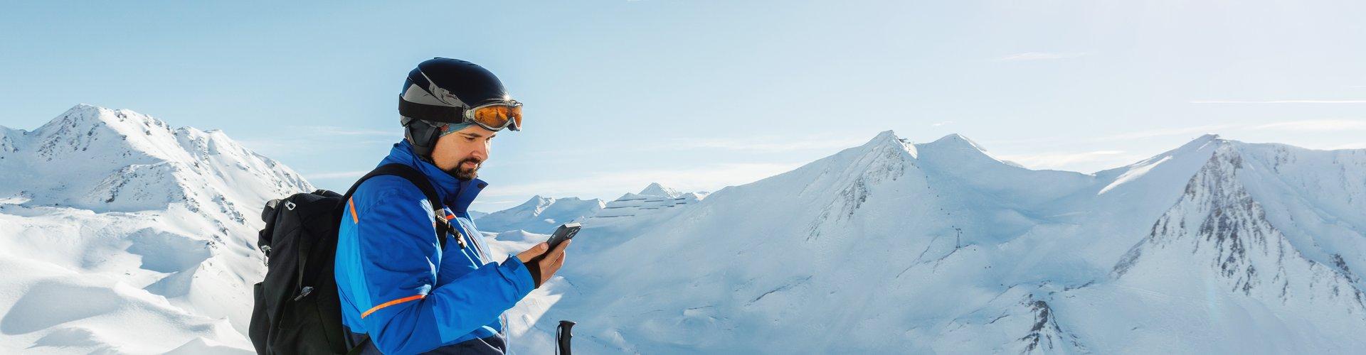 Ein Skifahrer steht auf einem schneebedeckten Gipfel und blickt auf sein Smartphone. Im Hintergrund erstreckt sich eine sonnige Berglandschaft mit schneebedeckten Gipfeln.